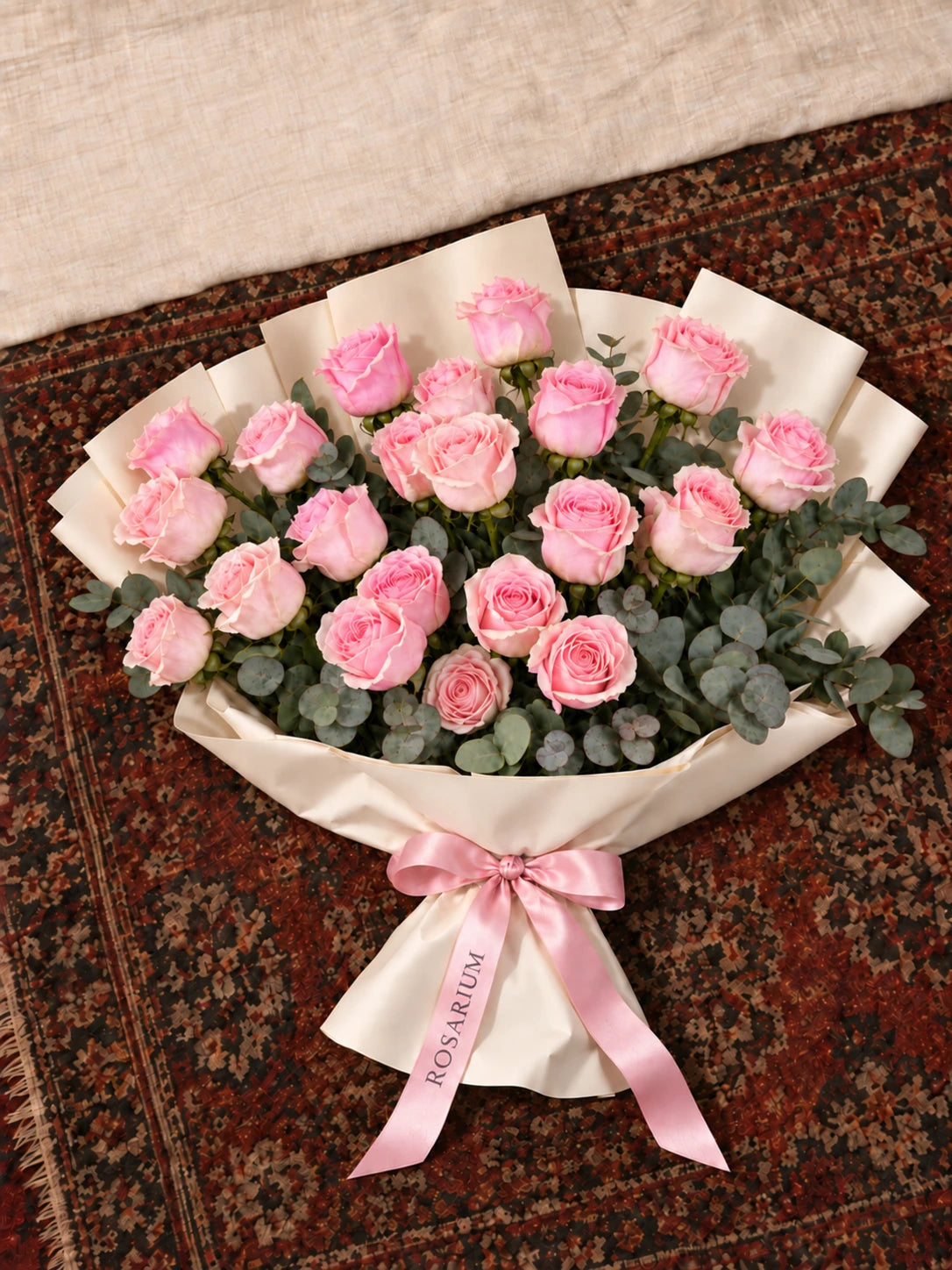 Pink roses and eucalyptus in ivory wrapping with a blush ribbon, photographed from above on a patterned carpet.
