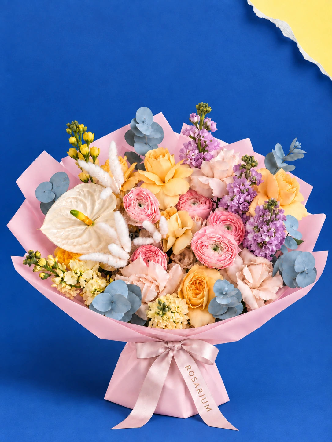 A bouquet of roses, anthuriums and ranunculus in shades of yellow, pink, cream and lilac, wrapped in pink paper.