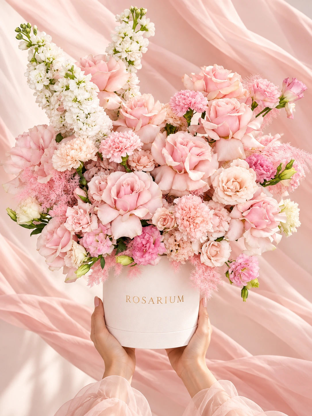 Roses and carnations in shades of pink and cream, arranged in an ivory round box. Also pictured: hands.