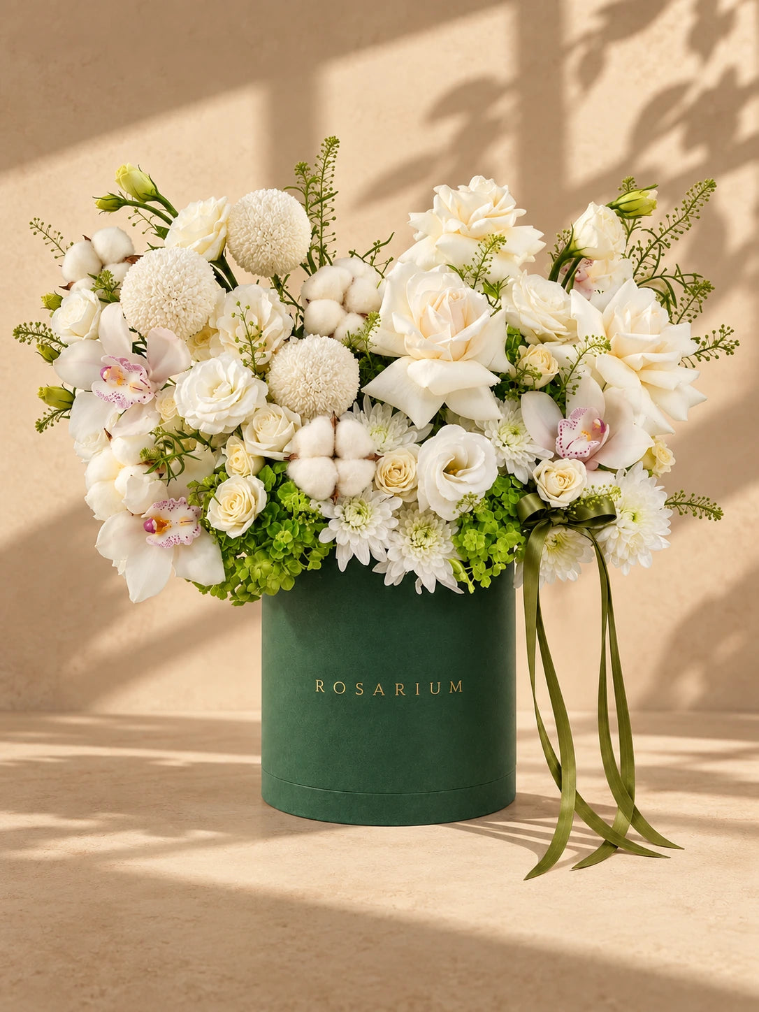 Ivory roses, white orchids, green hydrangea and round white blooms in a forest-green Rosarium box under warm window light.